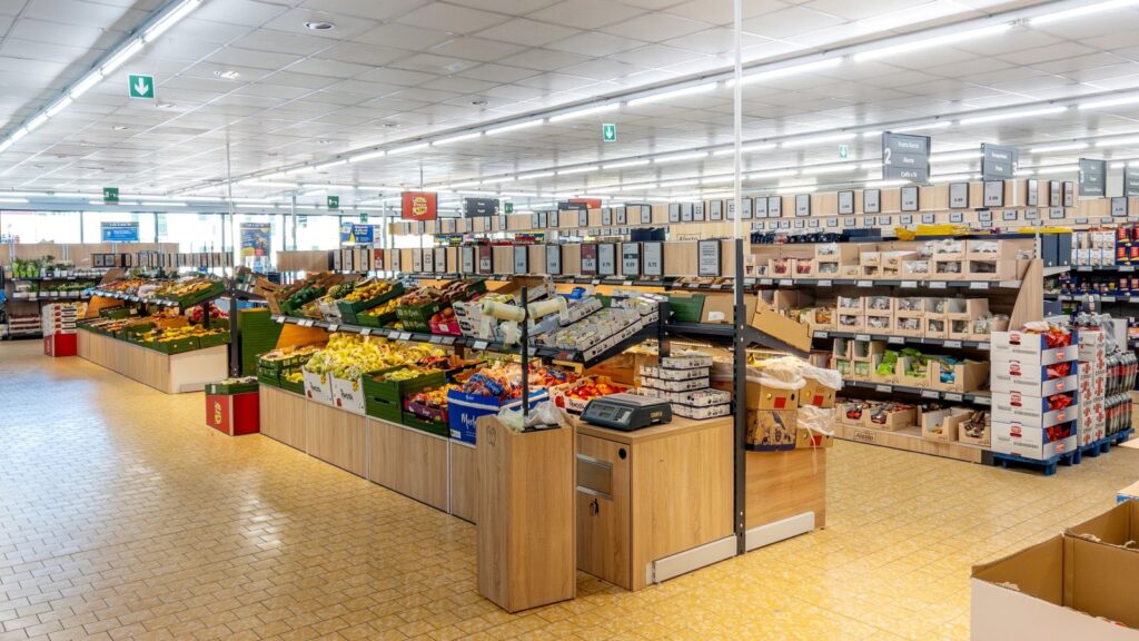A brightly lit grocery store interior with produce displays, boxed goods, and shelves stocked with various food items&mdash;including several things Americans ask for abroad; the store appears clean and organized with wide aisles.