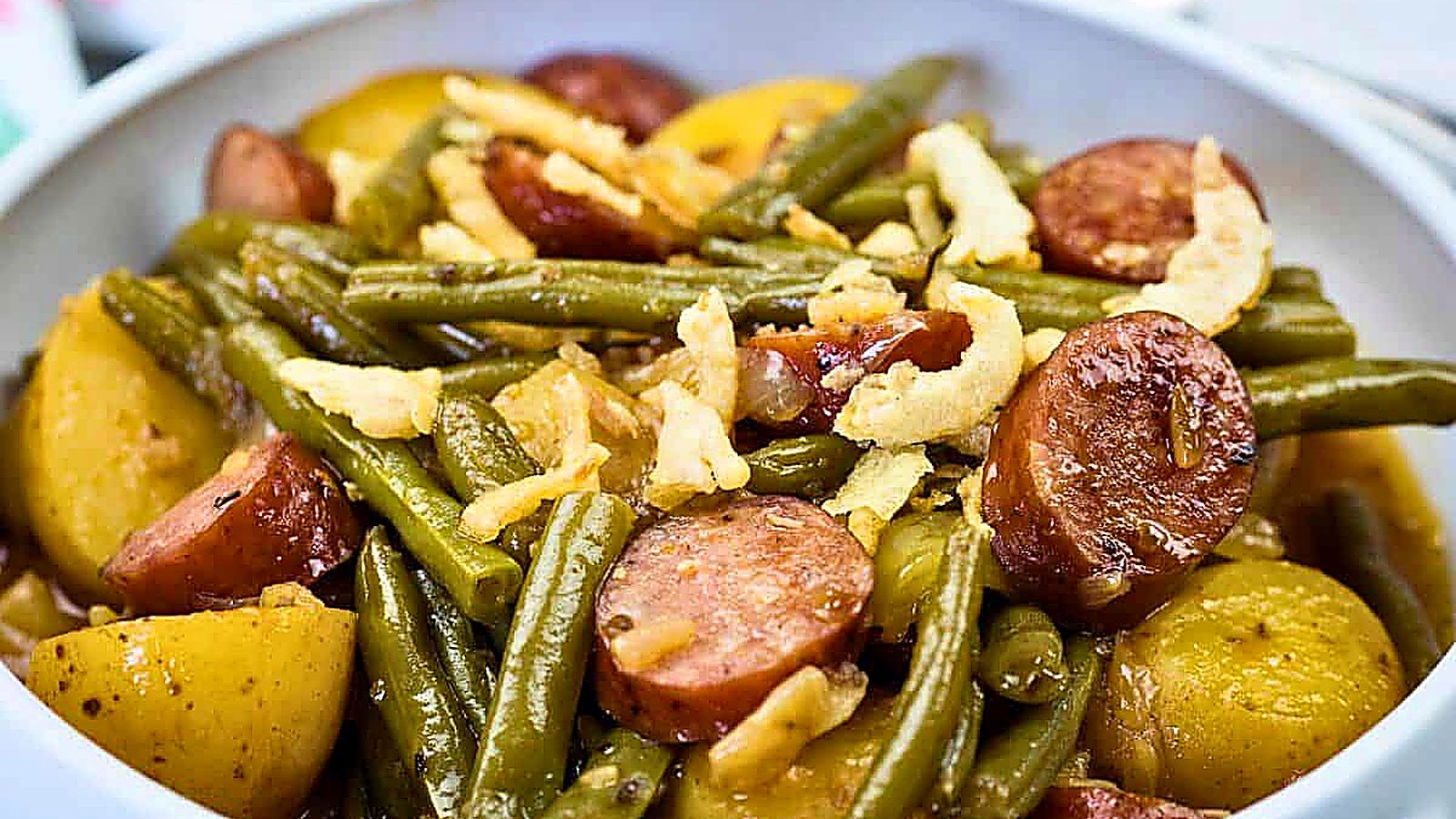 A bowl of Swamp Potatoes with cooked green beans, sliced sausage, and halved potatoes, topped with crispy fried onions.