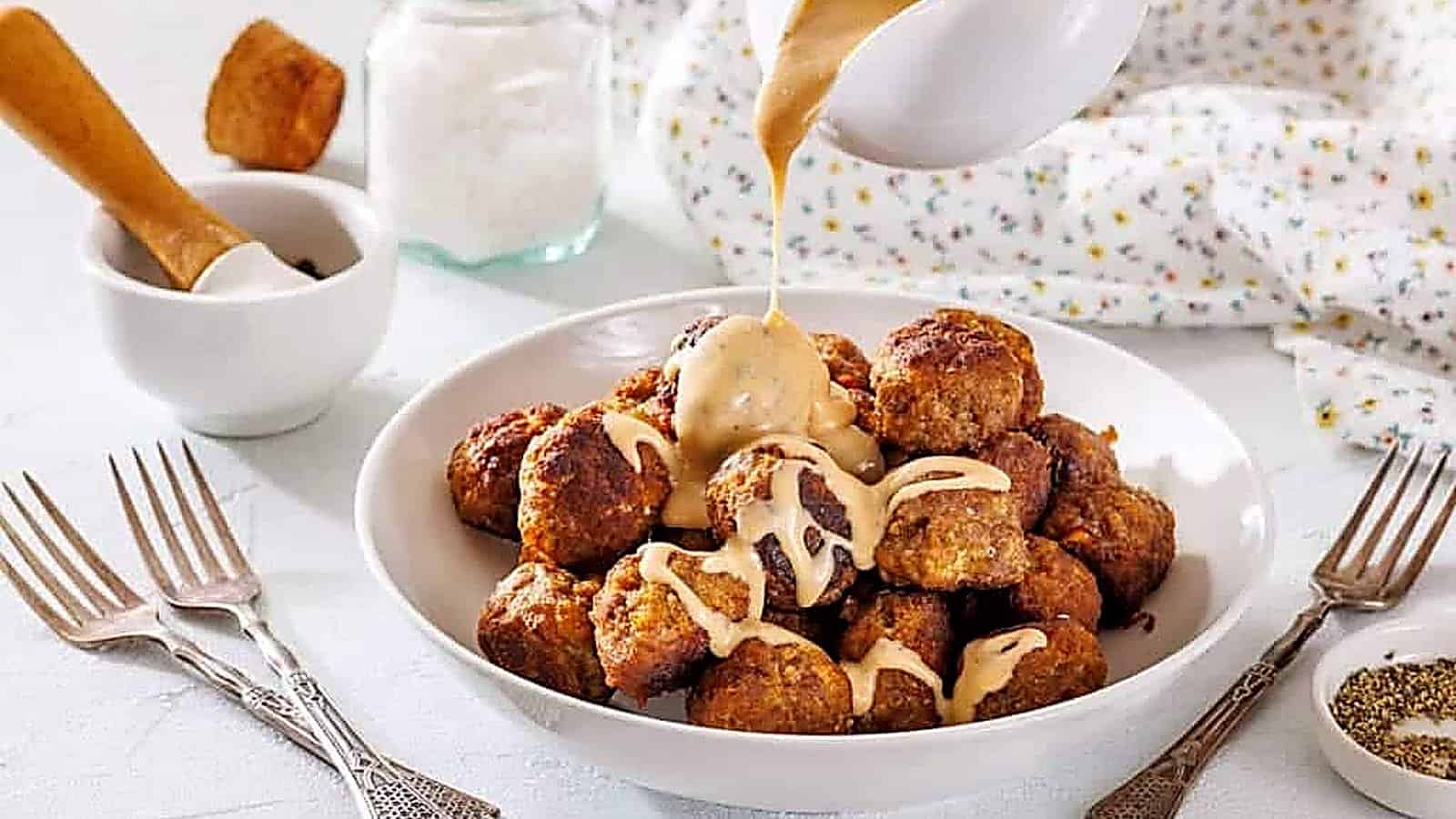 A bowl of meatballs is topped with creamy sauce being poured from a jug, surrounded by forks and seasoning jars on a white table.