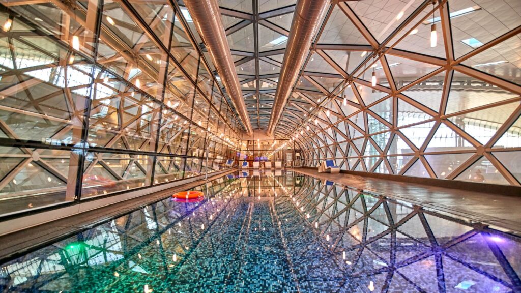 Indoor swimming pool with mosaic tile floor inside a modern glass and steel geodesic dome structure, with reflections on the water and ambient lighting&mdash;a luxurious feature you don&rsquo;t expect in an airport.