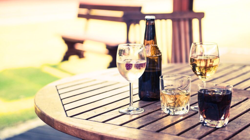Wooden table with four drinks: a wine glass with clear liquid and berries, a bottle of beer, a glass of whisky, and a glass of cola—perfect for relaxing outdoors while following BYOB rules in this inviting, blurred-background setting.