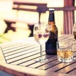 Wooden table with four drinks: a wine glass with clear liquid and berries, a bottle of beer, a glass of whisky, and a glass of cola—perfect for relaxing outdoors while following BYOB rules in this inviting, blurred-background setting.
