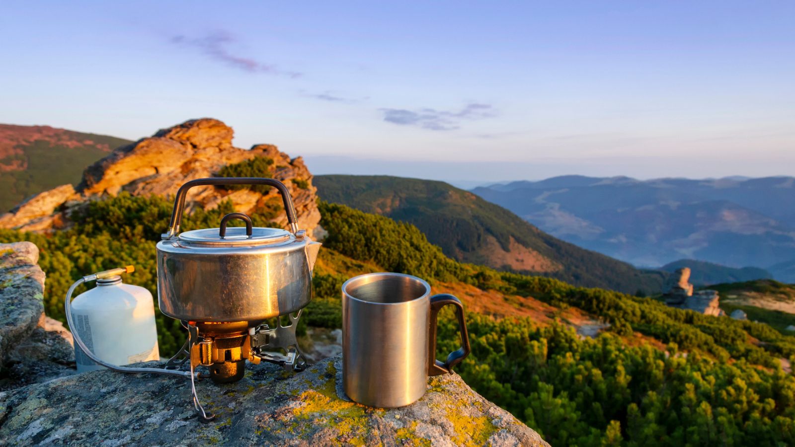 Camping stove, pot, and metal mug on a rocky surface with forested mountains and a clear sky in the background at sunrise or sunset.