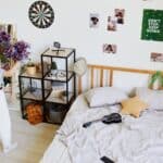 A person holding pom-poms stands near a shelf in a tidy bedroom with a dartboard, photos, and a “Don’t Stop. Keep Going.” poster on the wall. A ukulele lies on the bed.