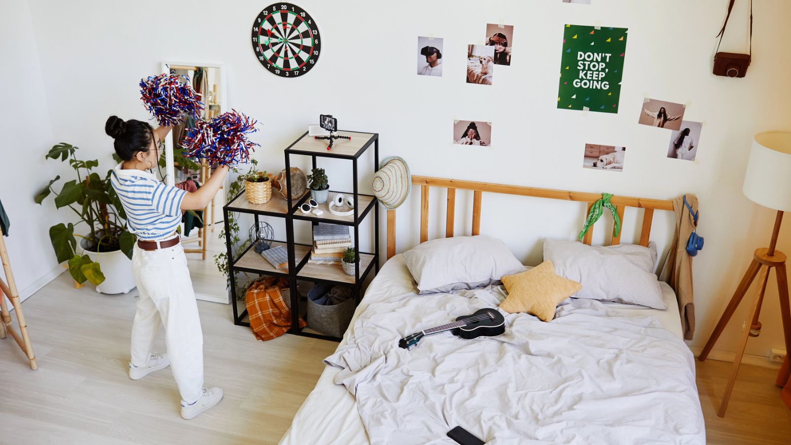 A person holding pom-poms stands near a shelf in a tidy bedroom with a dartboard, photos, and a “Don’t Stop. Keep Going.” poster on the wall. A ukulele lies on the bed.