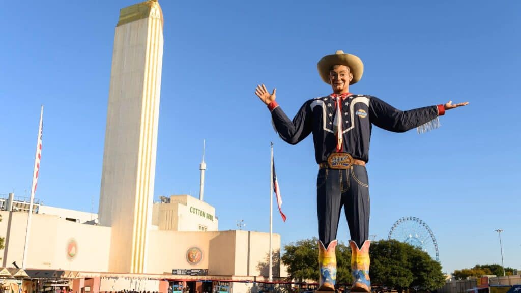 A large cowboy statue in western attire stands outdoors at the State Fair of Texas during state fair season, with a tall building and Ferris wheel in the background.