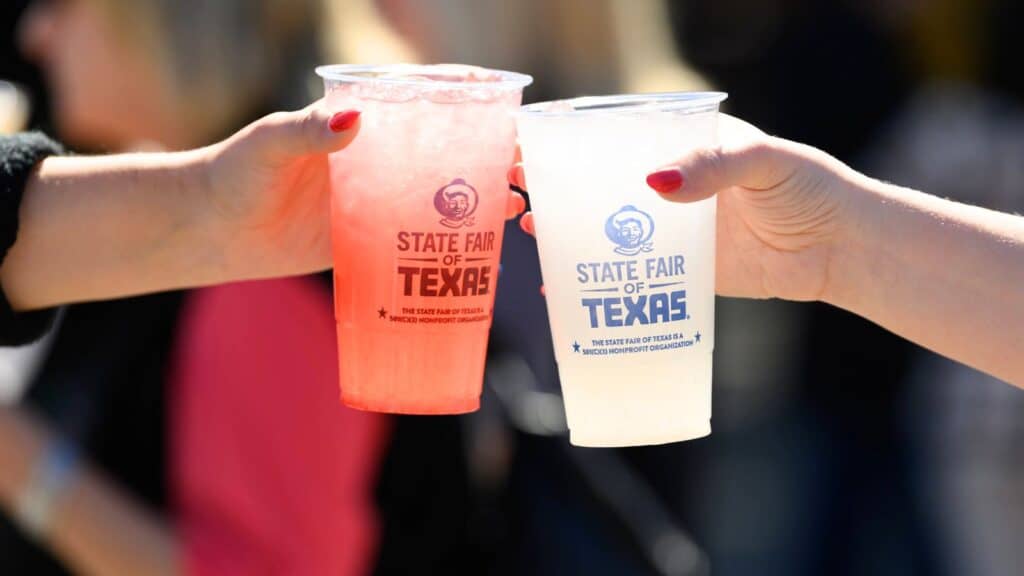 Two hands holding plastic cups with drinks, labeled &ldquo;State Fair of Texas,&rdquo; clink cups together at an outdoor event.