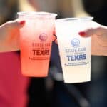 Two hands holding plastic cups with drinks, labeled “State Fair of Texas,” clink cups together at an outdoor event.
