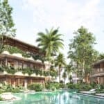 A tropical resort features two-story buildings with balconies, lush greenery, palm trees, and a central pool surrounded by lounge chairs under a partly cloudy sky.