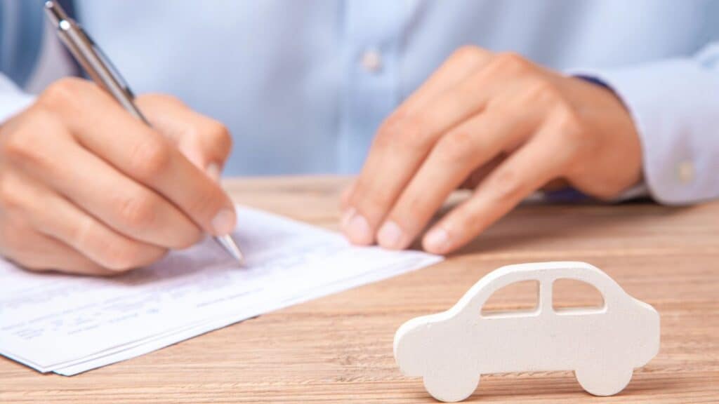 A person fills out paperwork at a desk with a wooden car model in the foreground, highlighting how unexpected rental car fees catch travelers during the booking process.