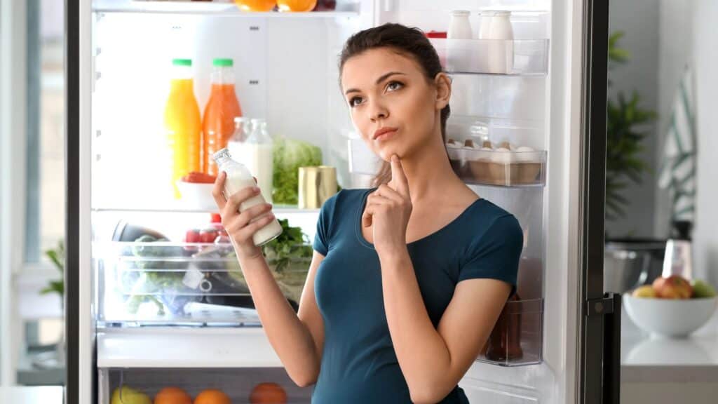 Woman stands in front of an open refrigerator, holding a bottle of milk and looking thoughtful, surrounded by various foods you are refrigerating and drink items.