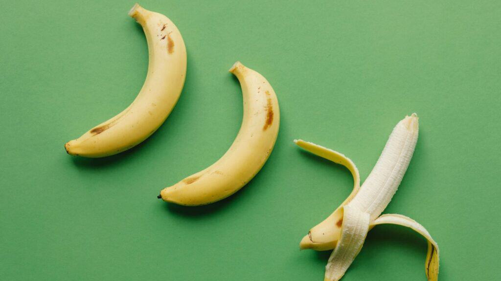 Two whole bananas and one half-peeled banana are arranged on a plain green background, creating a vibrant celebration perfect for National Banana Lovers Day.