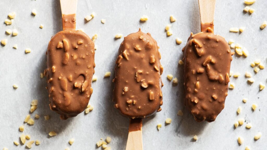 Three chocolate-coated ice cream bars with nuts on wooden sticks, arranged in a row on a gray surface with scattered nut pieces&mdash;classic ice cream truck treats perfect for satisfying your sweet tooth.
