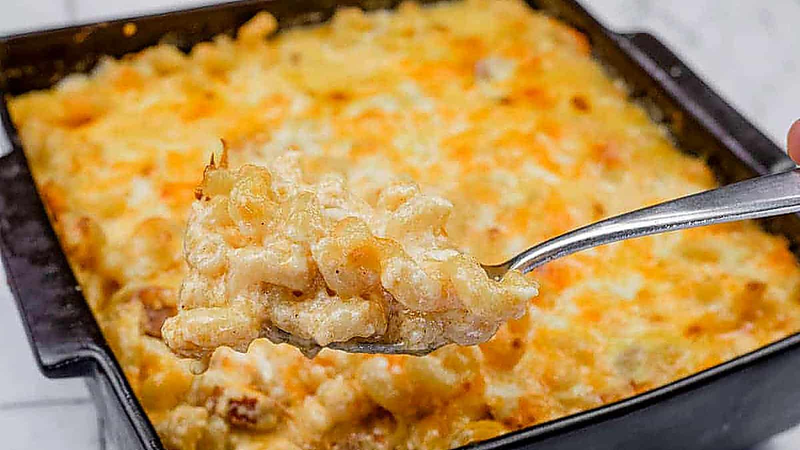 A spoonful of Tini&rsquo;s Mac & Cheese with a Twist is held above a square baking dish filled with the same dish.