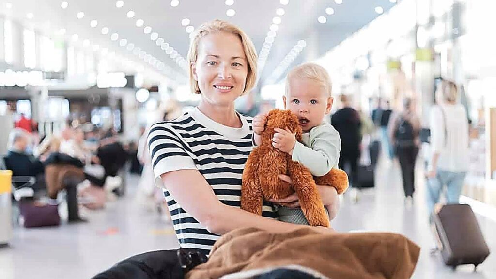 A woman holding a baby and a stuffed toy stands in a busy airport terminal with people and luggage in the background.