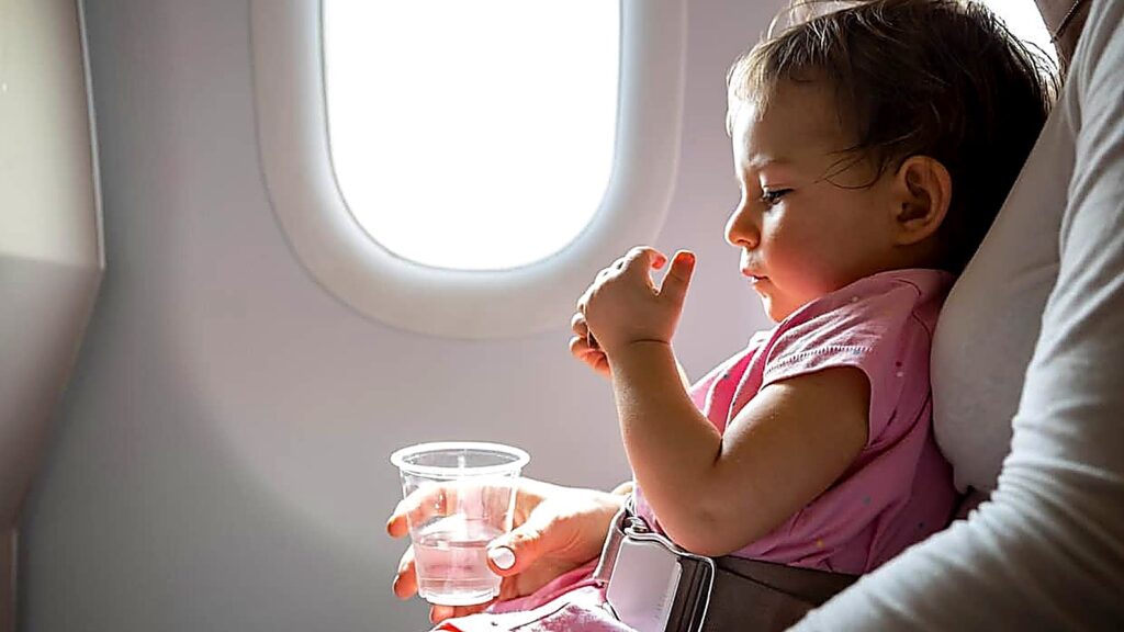 A toddler in a pink shirt sits on an adult&rsquo;s lap on an airplane, holding a cup of water, with a window and seatbelt visible.