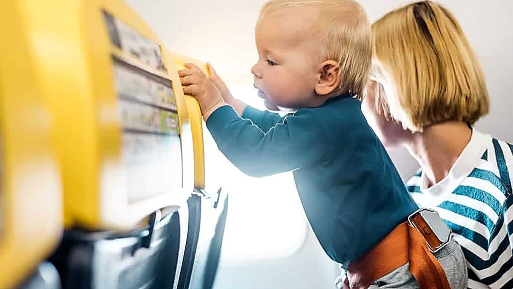 A baby stands on an airplane seat, holding onto the backrest, while an adult with short blond hair sits beside them.