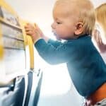 A baby stands on an airplane seat, holding onto the backrest, while an adult with short blond hair sits beside them.