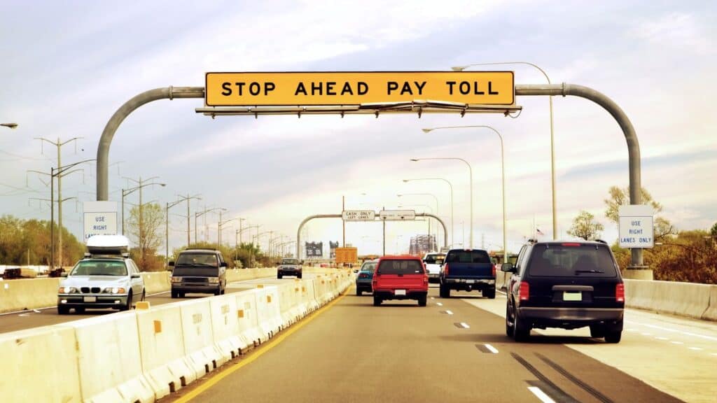 Vehicles drive on a highway toward a yellow overhead sign that reads &ldquo;STOP AHEAD PAY TOLL.&rdquo; Concrete barriers separate lanes and cars are visible in both directions, reminding travelers to watch for unexpected costs like rental car fees that often catch travelers by surprise.