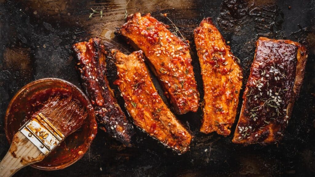 Five glazed pork ribs with sesame seeds on a dark tray, next to a bowl of sauce with a basting brush&mdash;a delicious example of American foods with foreign roots.