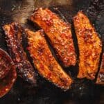 Five glazed pork ribs with sesame seeds on a dark tray, next to a bowl of sauce with a basting brush—a delicious example of American foods with foreign roots.