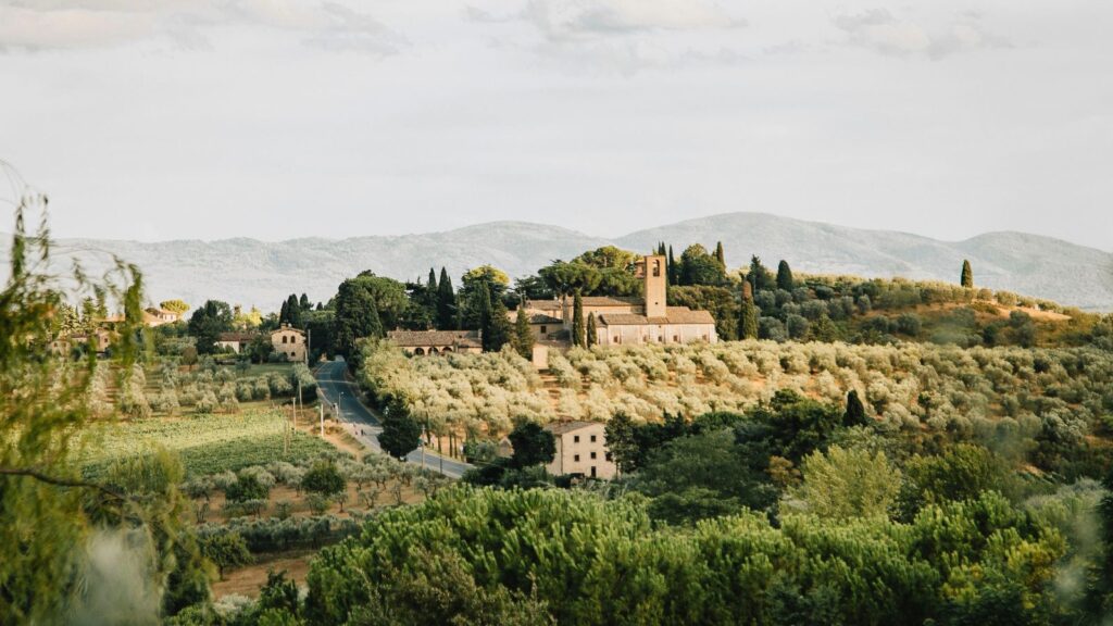A rural landscape with a stone church and scattered buildings among green trees and fields, set against distant hills under a cloudy sky&mdash;an idyllic scene perfect for Routescape luxury travel seekers in search of serene elegance and timeless countryside charm.