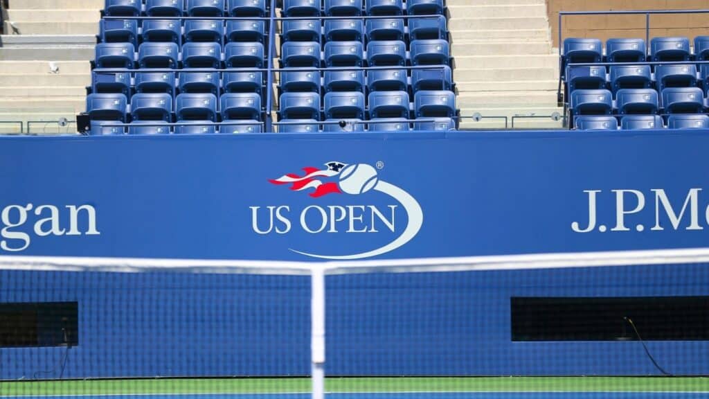 Empty stadium seats behind a blue barrier with the US Open tennis tournament logo and sponsor names, viewed from across a tennis net.