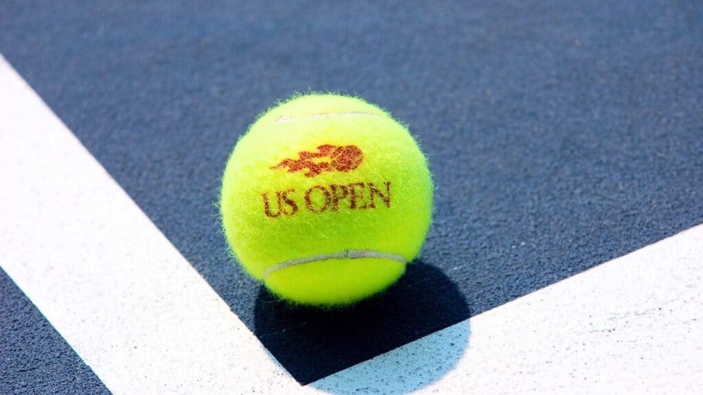 A yellow tennis ball with a US Open logo rests on a blue and white tennis court near the intersection of two white lines.