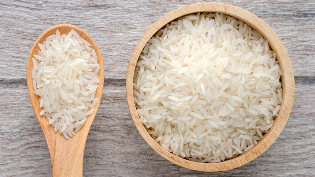 A wooden bowl filled with uncooked white rice sits next to a wooden spoon holding a small portion of rice on a light wooden surface.