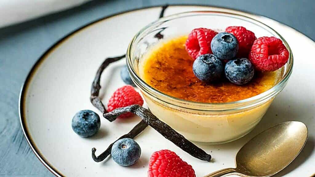 A glass bowl of cr&egrave;me br&ucirc;l&eacute;e topped with fresh raspberries and blueberries, served on a plate with a gold spoon and vanilla beans.