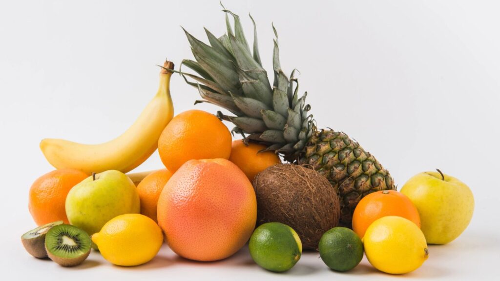Assorted fresh fruits, including pineapple, bananas, oranges, apples, grapefruit, lemons, limes, a coconut, and a sliced kiwi&mdash;some of which are foods US customs may confiscate&mdash;are arranged on a white background.