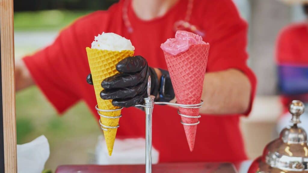 A person wearing a red shirt and black glove holds yellow and pink waffle cones with scoops of ice cream at an outdoor stand.