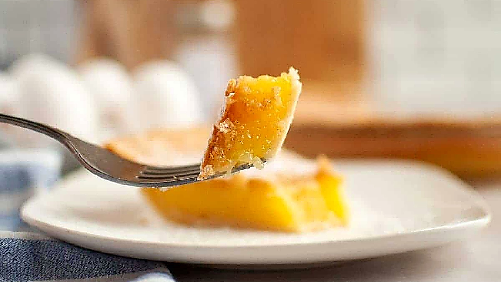 A fork holds a bite of Vinegar pie above a plate with a larger slice, with eggs and kitchen items blurred in the background.