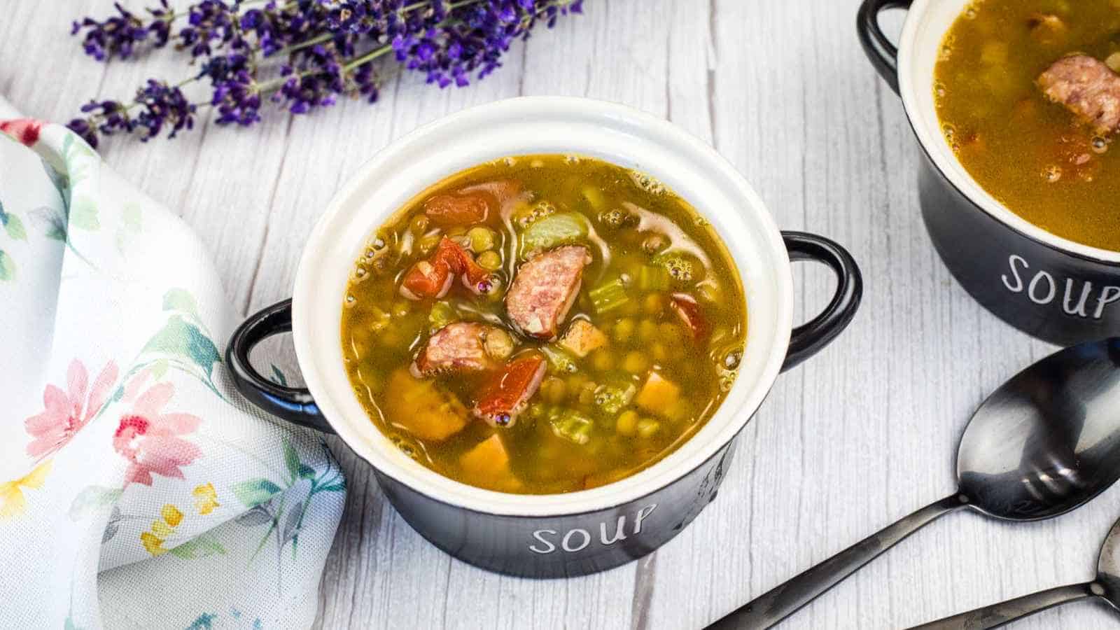 A bowl of lentil soup sits on a light wooden table next to a floral napkin, a sprig of lavender, and a black spoon.