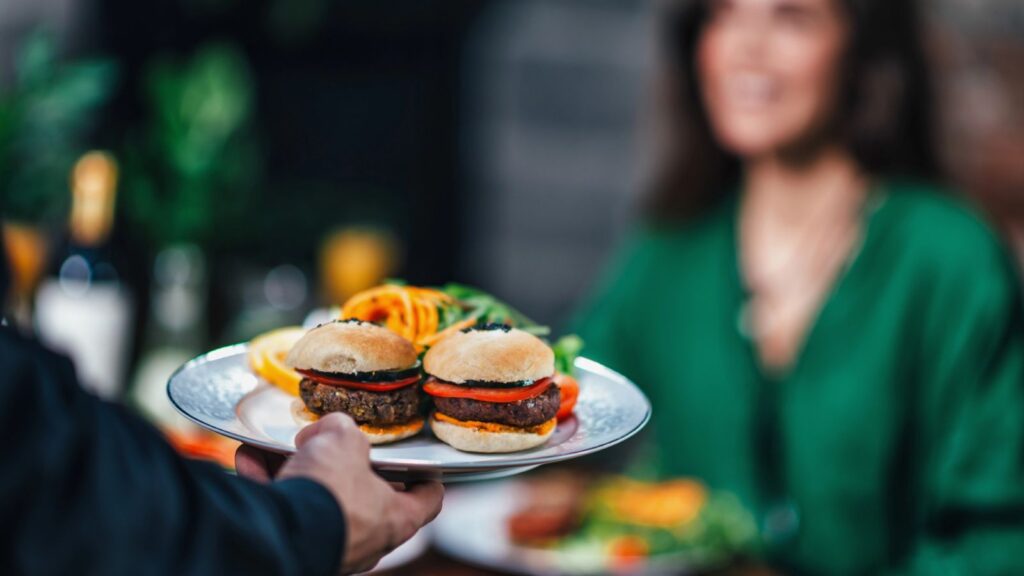 A server presents a plate with two small burgers and garnish to a seated woman in a green top at a restaurant table, highlighting the unique flavors that regional chains rarely cross state lines to offer.