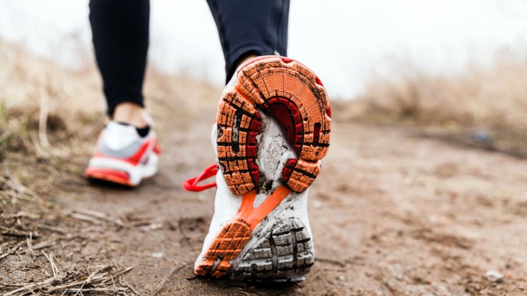 Close-up view of a person walking on a dirt path, focusing on the sole of a white and orange running shoe. The background is blurred and shows dry grass.