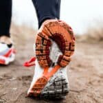 Close-up view of a person walking on a dirt path, focusing on the sole of a white and orange running shoe. The background is blurred and shows dry grass.