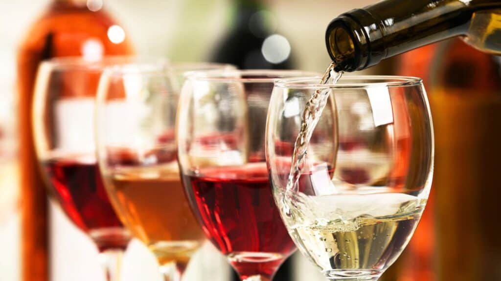 A bottle pours white wine into a glass, with other glasses of red, ros&eacute;, and white wine lined up in the background.