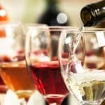 A bottle pours white wine into a glass, with other glasses of red, rosé, and white wine lined up in the background.
