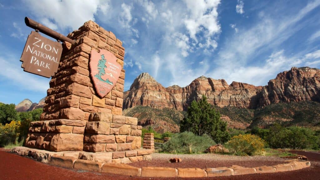 A stone entrance sign for Zion National Park stands in front of rugged red rock mountains under a blue sky with scattered clouds, welcoming visitors to one of the many US national parks that require reservation for entry.