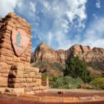 A stone entrance sign for Zion National Park stands in front of rugged red rock mountains under a blue sky with scattered clouds, welcoming visitors to one of the many US national parks that require reservation for entry.