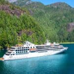 A white cruise ship sails through calm, blue water surrounded by dense green forest and mountains under a clear sky.