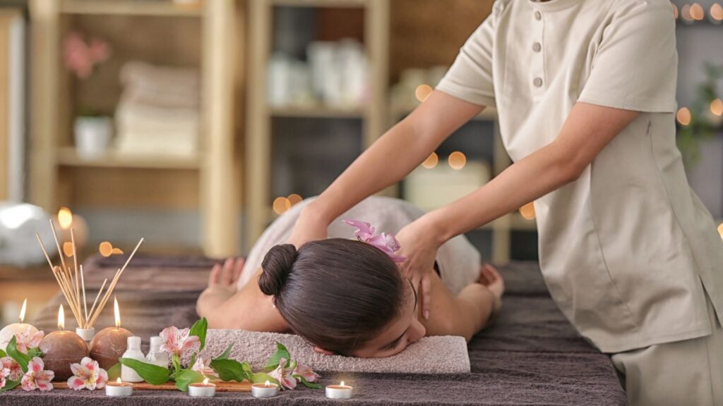 Person receiving a back massage in a spa setting you don&rsquo;t expect in airport, surrounded by candles, flowers, and incense on a table.