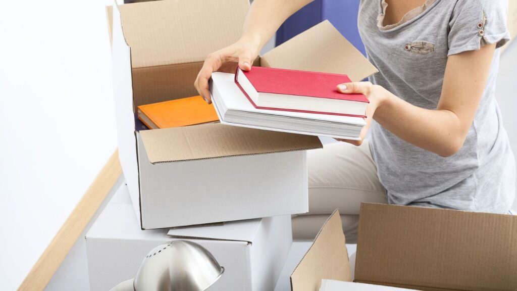 A person wearing a gray shirt packs books into a cardboard box, with other boxes and a desk lamp nearby, preparing for an early college move-in.