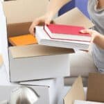 A person wearing a gray shirt packs books into a cardboard box, with other boxes and a desk lamp nearby, preparing for an early college move-in.