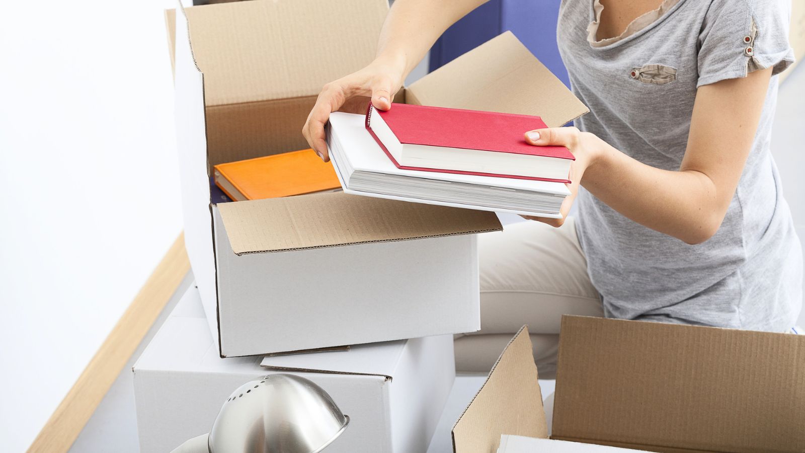 A person wearing a gray shirt packs books into a cardboard box, with other boxes and a desk lamp nearby, preparing for an early college move-in.