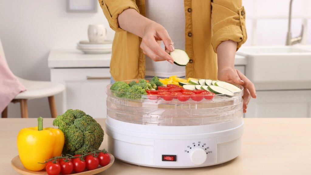 Person placing sliced zucchini&mdash;one of the best foods to dehydrate&mdash;into a food dehydrator filled with assorted vegetables, including broccoli, tomatoes, and bell peppers, on a kitchen counter.