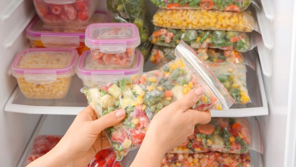 Hands holding a plastic bag of mixed frozen vegetables in front of an American freezer filled with various frozen vegetables, bags, containers, and convenient frozen dinner options.