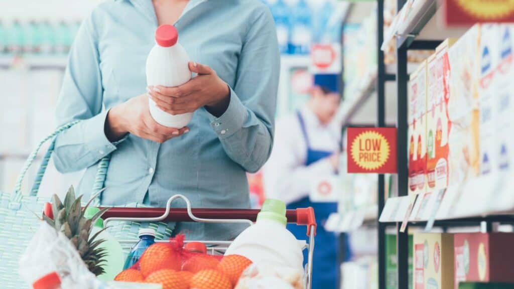 A person holding a bottle in front of grocery shelves, with a basket of fruit and groceries in a cart, pauses beneath a &ldquo;Low Price&rdquo; sign&mdash;proof that food labeling terms aren&rsquo;t simple, as every choice requires careful attention.