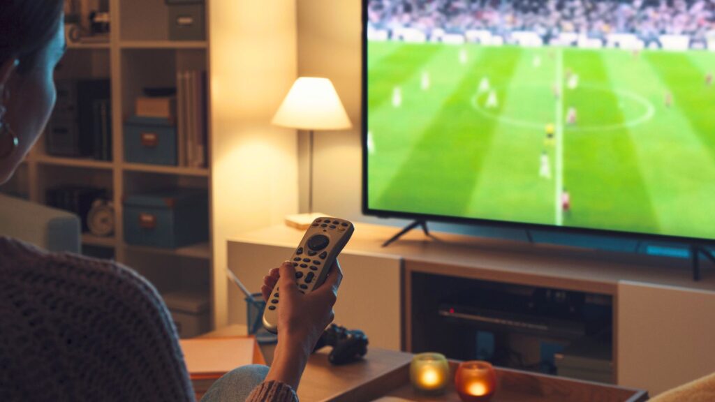 Person holding a remote control and watching a free World Cup stream on a flat-screen TV in a cozy living room with candles and shelves in the background.
